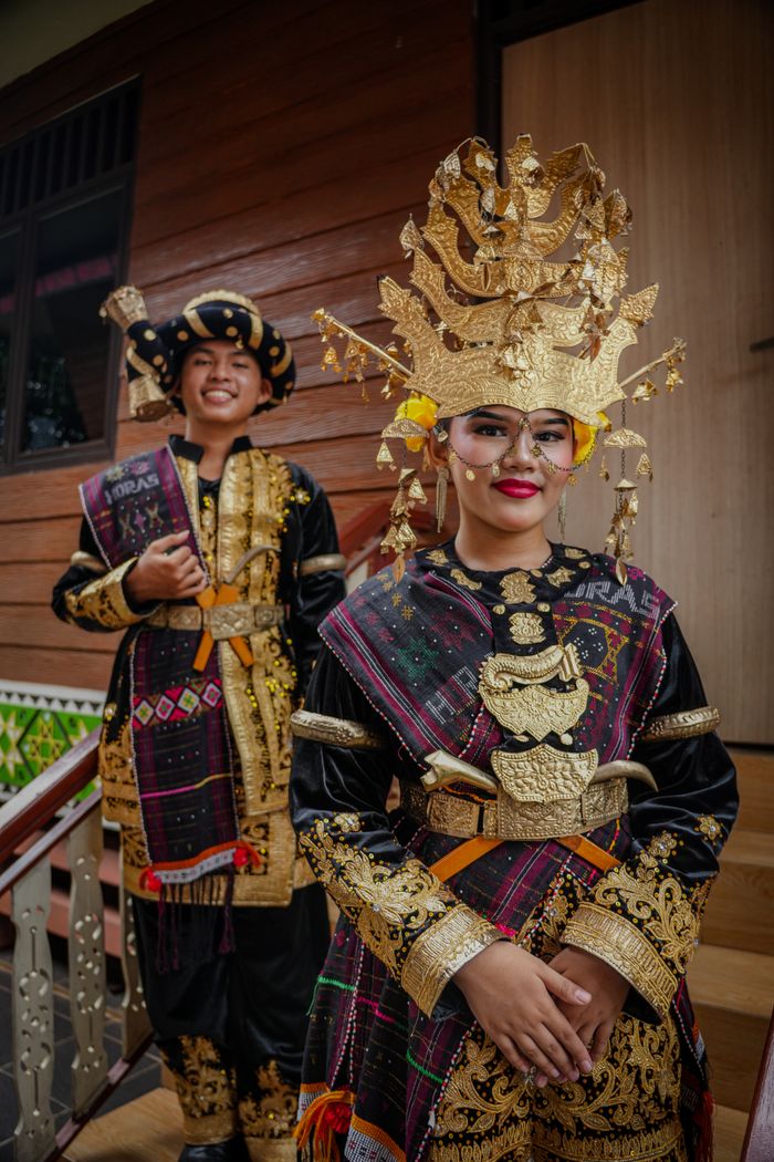 Two people in traditional Indonesian wedding attire on wooden stairs, showcasing cultural heritage.